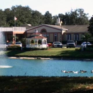 A peaceful scene featuring a building by a lake with a fountain, ducks swimming, and a gazebo surrounded by trees.