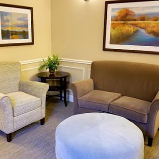 A cozy seating area featuring a light-colored armchair, a brown sofa, an ottoman, and decorative artwork.