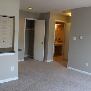 An empty apartment room with a carpeted floor and neutral-colored walls, featuring a closet and open doorway.