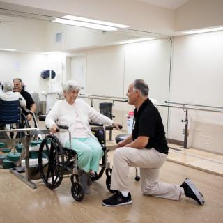 A physical therapist kneels beside an elderly person in a wheelchair, providing support and care in a rehabilitation setting.