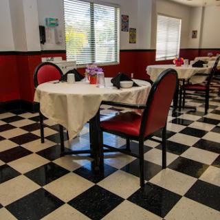 A cozy dining area featuring tables draped in cream-colored tablecloths and red chairs, illuminated by natural light.