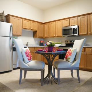 A cozy kitchen with a small dining area featuring a round table and two cushioned chairs, surrounded by wooden cabinets and stainless steel appliances.