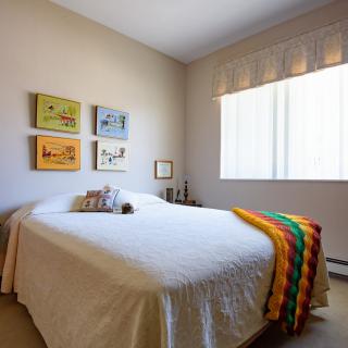 A cozy bedroom with a neatly made bed, colorful artwork on the wall, and a bright window allowing natural light in.