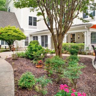 A beautifully landscaped garden with colorful flowers and a tree, surrounded by a pathway in front of a building.