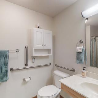A clean bathroom featuring a toilet, sink, and mirror with accessible grab bars and towel racks.