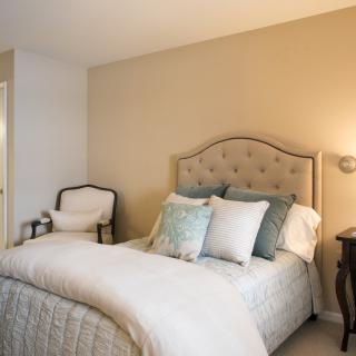 A cozy and elegantly styled bedroom with a bed adorned with multiple pillows, a chair in the corner, and a lamp on a bedside table.