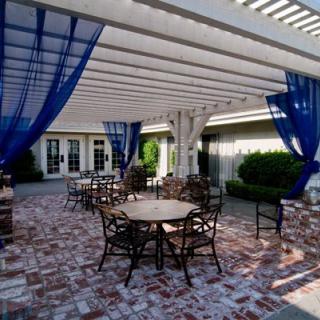 A charming outdoor patio area featuring tables and chairs under a pergola with blue curtains, set on a brick floor.