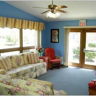A cozy living room with floral sofas, an armchair, and a ceiling fan, illuminated by natural light through large windows.