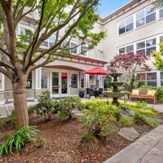 A peaceful courtyard with lush greenery, outdoor seating, and a fountain in front of a multi-story building.