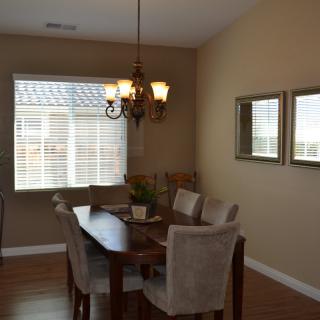 A cozy dining room with a wooden table, upholstered chairs, and a chandelier, featuring large windows and wall decor.