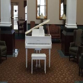 A white grand piano is placed in the center of a lobby with chairs, columns, and a patterned carpet.