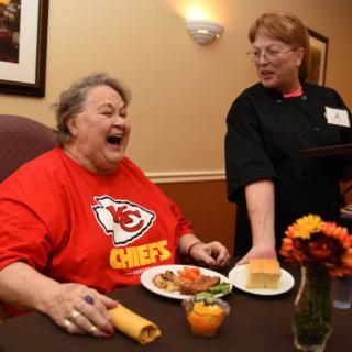 An elderly person wearing a Kansas City Chiefs shirt shares a joyful moment with a staff member during mealtime at a nursing home.