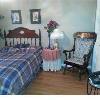 A cozy bedroom with a plaid bedspread, a wooden chair, and a small nightstand with a lamp and flowers.