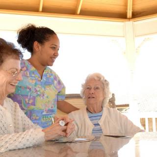 A caregiver interacting with two elderly women at an outdoor table.