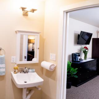 A view of a bathroom with a sink and mirror, adjacent to a living area featuring a TV and decorative elements.
