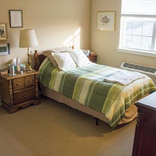 A cozy bedroom with a bed covered in a green blanket, accompanied by a nightstand and lamp near the window.
