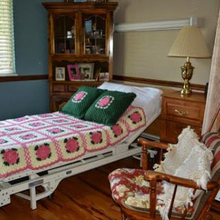 A cozy bedroom featuring a hospital bed adorned with a colorful quilt and decorative pillows, alongside an armchair and wooden furniture.