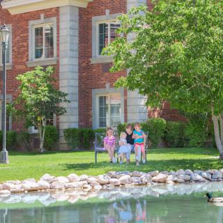 A family sitting on a bench by a pond in a park setting with a brick building in the background.