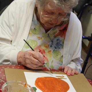An elderly woman engaged in painting a picture of a pumpkin.