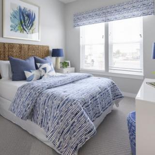 A modern bedroom with a blue and white color scheme, featuring a bed adorned with patterned bedding and decorative pillows. The room is brightened by natural light from the large window, complemented by artwork on the wall.