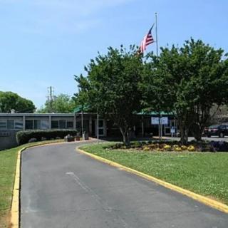 The image shows the entrance of a school building with a driveway, surrounded by trees and displaying an American flag on a flagpole.