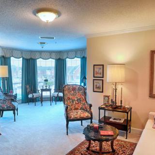 A cozy living room with traditional decor, featuring armchairs, a carpet, and large windows with curtains.