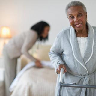 An elderly woman using a walker with a caregiver making the bed in the background.