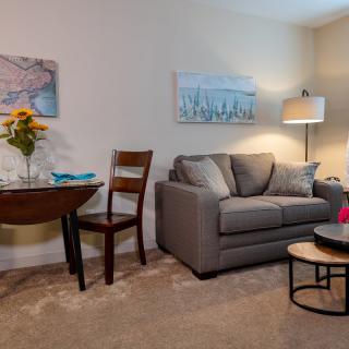 A cozy living room with a gray sofa, wooden chair, and round coffee table, decorated with artwork and a standing lamp.