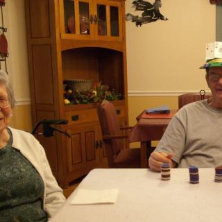 An elderly couple sitting at a dining table, smiling and enjoying a casual moment indoors.
