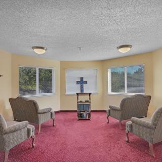 A small room with upholstered chairs, a red carpet, and a cross on a stand in front of windows.
