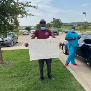 Two individuals in a parking lot, one holding a sign and the other wearing scrubs.