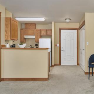 A cozy kitchen area in an apartment with wooden cabinets and a carpeted floor, featuring an open layout.