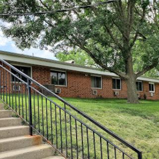 A view of a brick residential building with stairs leading up to it, surrounded by lush green grass and trees.