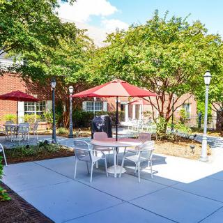 A sunny outdoor patio area with tables, chairs, and red umbrellas surrounded by trees.