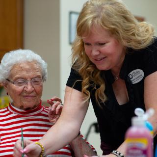 A caregiver assisting an elderly woman with a creative activity.