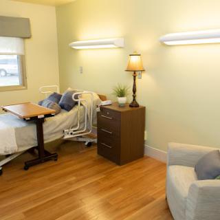 A well-lit hospital room featuring a bed, nightstand with lamp, and a chair.