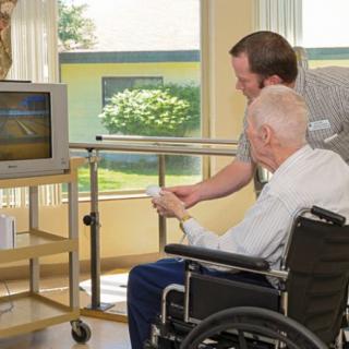 An elderly man in a wheelchair plays a video game with assistance in a nursing home setting.