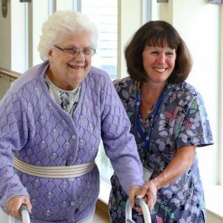 A caregiver is assisting a smiling elderly woman using a walker.
