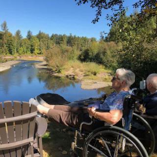 Two people in wheelchairs enjoy a peaceful view of the river and surrounding forest.