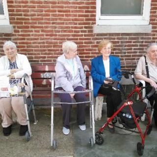 Four elderly women sitting on a bench with walkers in front of a brick wall.