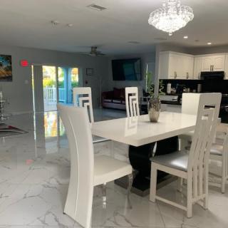 A modern dining area with white furniture and a chandelier, opening into the kitchen.