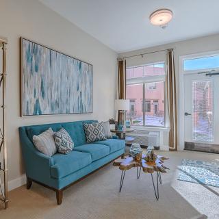 A cozy living room with a teal sofa, abstract artwork on the wall, and natural light streaming in through large windows.