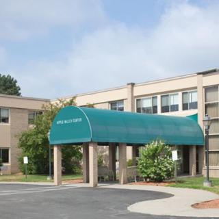 An entrance with a green awning leading into a building surrounded by greenery under a cloudy sky.