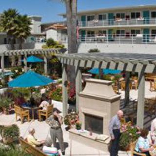 A lively outdoor patio area with people seated at tables under blue umbrellas, next to a fireplace and surrounded by a multi-story building.