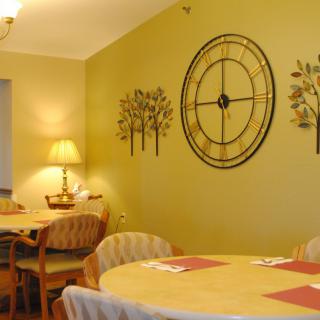 A cozy dining room with round tables, chairs, a large wall clock, and warm lighting.