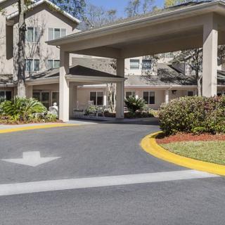 The image shows the entrance and driveway of a residential building, surrounded by landscaped gardens and trees.