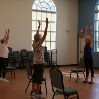 A group of women participating in a yoga class with chairs in a sunlit room.