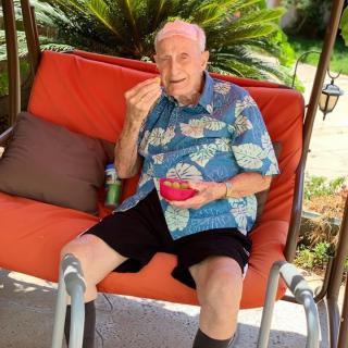 An elderly man is sitting on an outdoor swing chair, enjoying a snack from a bowl.