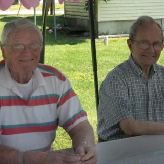 Two elderly men sitting at a table outdoors, smiling.