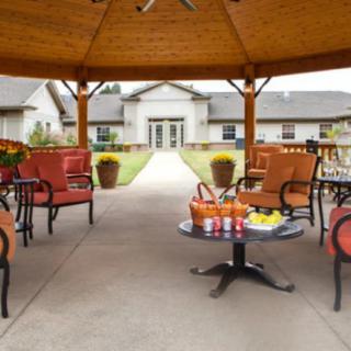 A cozy outdoor seating area under a gazebo, featuring comfortable chairs and a table with refreshments set against a backdrop of landscaped gardens.